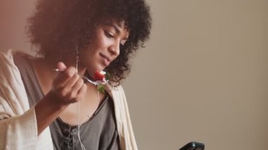 Memnuniyet verici konsantre olmuş Afrikalı kadın evde cep telefonu kullanıyor ve kahvaltı yaparken kulaklıkla müzik dinliyor.