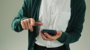 Stüdyoda gri bir duvarın üzerinde duran akıllı telefonuyla yazı yazan genç bir adamın kırpılmış görüntüsü.