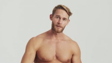 Pozitif yarı çıplak sarışın bir adam stüdyodaki gri bir arka planda tek başına barış jesti yapıyor.
