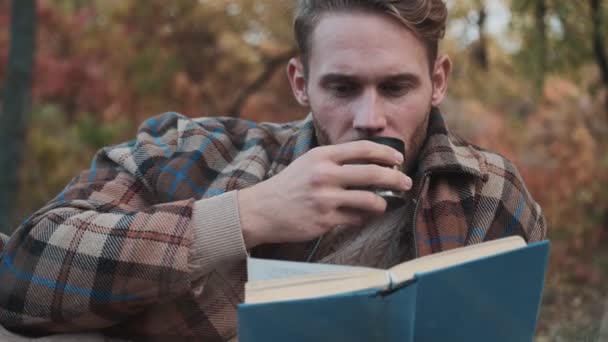 一个专心致志的男人坐在秋天的公园里喝着热茶 一边看书 — 图库视频影像
