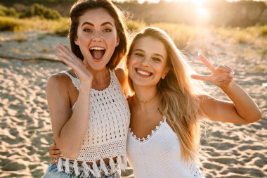 İki beyaz kadın, dışarıda yürürken gülümsüyor ve barış işareti yapıyor.