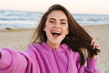 Beyaz tenli güzel bir kadın dışarıda yürürken gülümsüyor ve selfie çekiyor.