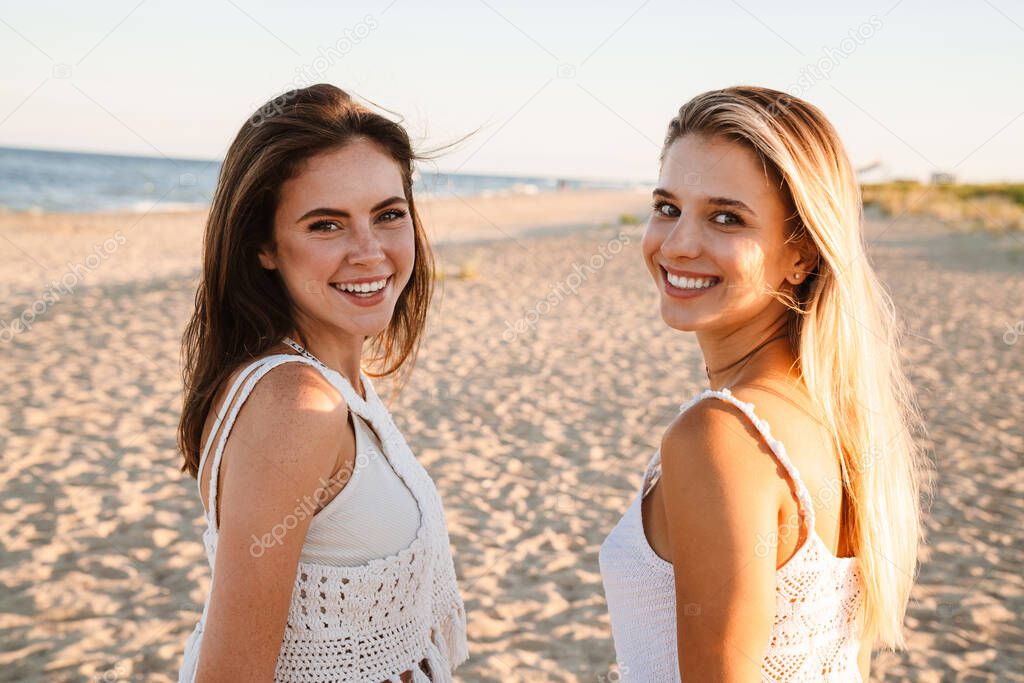Two young caucasian happy women smiling and looking at camera while walking on beach