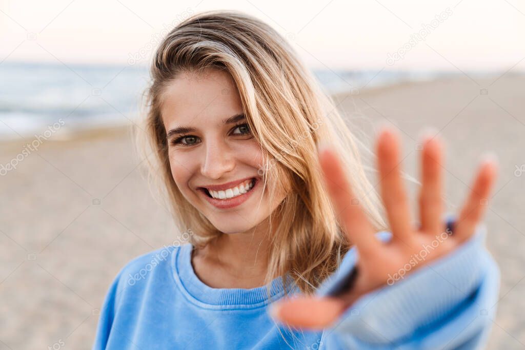 Young caucasian blonde woman smiling and gesturing her palm at camera while walking outdoors
