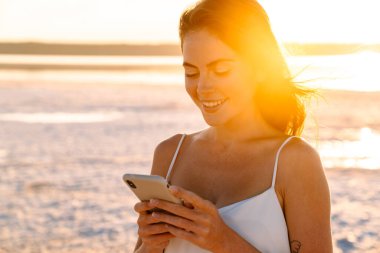 Sahilde yürüyen ve cep telefonu kullanan gülümseyen genç bir kız resmi