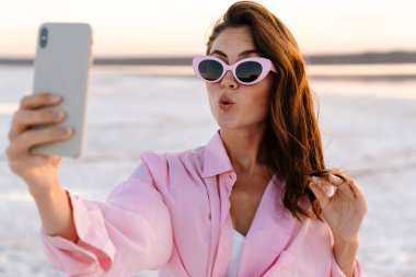Güneş gözlüklü genç bir kızın dışarıda sahilde telefonla selfie çekerken çekilmiş görüntüsü.