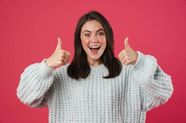 Heyecanlı güzel bir kızın pembe arka planda izole bir şekilde başparmağını kameraya göstermesi.