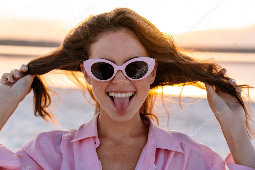 Image of a positive young pretty girl in sunglasses posing outdoors at the beach and showing tongue