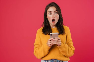 Şok olmuş esmer kız görüntüsü. Pembe arka planda izole edilmiş cep telefonu pozu ve kullanımı.