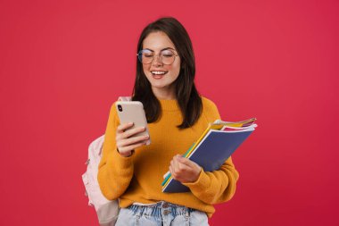 Pembe arka planda izole edilmiş egzersiz kitaplarıyla poz verirken cep telefonu kullanan mutlu öğrenci görüntüsü