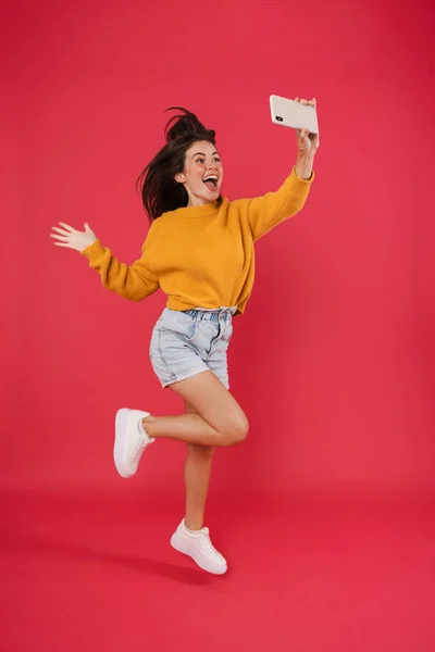 照片中 兴奋的黑发女孩在手机上拍下自己的照片 同时在粉色背景下跳下与外界隔绝 — 图库照片