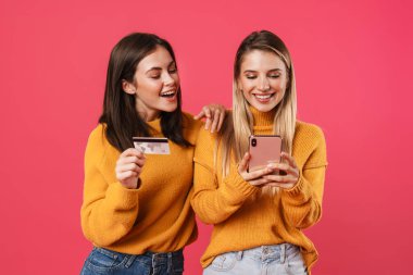 Cep telefonu ve kredi kartıyla online alışveriş yapan iki mutlu genç kız arkadaş pembe duvar arkasında izole edilmiş.
