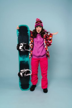 Şaşırdığını ifade eden kafası karışmış sporcu kadın mavi duvarın üzerinde izole edilmiş bir snowboard ile poz veriyor.