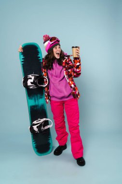 Güzel, heyecanlı sporcu kadın mavi duvarın üstünde kar kaykayıyla poz verirken kahve içiyor.