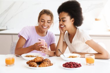 Güzel, neşeli, çok kültürlü kızlar ev mutfağında kahvaltı ederken cep telefonu kullanıyorlar.