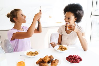 Güzel, neşeli, çok kültürlü kızlar cep telefonuyla fotoğraf çektirirken ev mutfağında beraber kahvaltı ediyorlar.