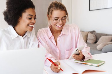 Gülümseyen çok kültürlü öğrenciler cep telefonlarını evde birlikte ev ödevi yaparken kullanıyorlar.