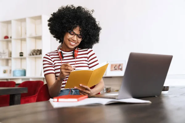 アフリカ系アメリカ人の学生の女の子を笑顔ノートパソコンで宿題をしながらテーブルに座って屋内 — ストック写真