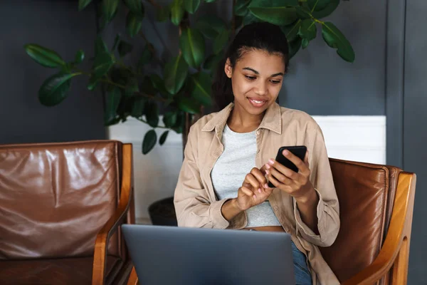 坐在咖啡店里用笔记本电脑和手机工作的非洲裔美国女人笑着 — 图库照片
