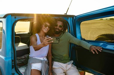 Genç çift arabada otururken akıllı telefon kullanarak selfie çekiyor.