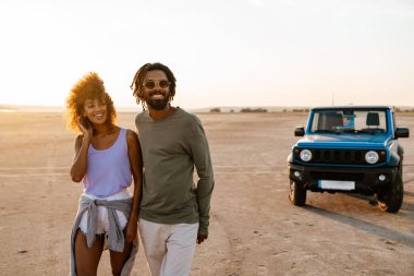 Çölde arabayla gezerken kucaklaşan ve yürüyen neşeli Afro-Amerikan çifti.