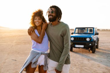 Çölde arabayla gezerken kucaklaşan ve yürüyen neşeli Afro-Amerikan çifti.