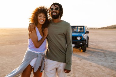 Çölde arabayla gezerken kucaklaşan ve yürüyen neşeli Afro-Amerikan çifti.
