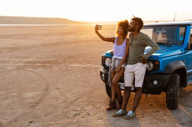 Çölde arabayla seyahat ederken cep telefonundan selfie çeken Afro-Amerikan çifti.