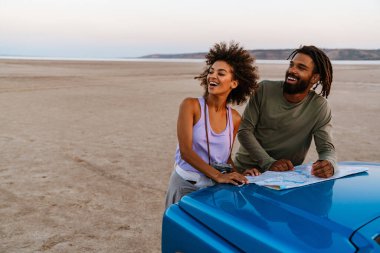 Çölde arabayla seyahat ederken haritayı inceleyen neşeli Afrikalı Amerikalı çift.