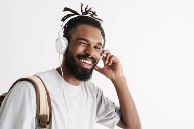 Beyaz duvarda izole edilmiş kulaklıklarla müzik dinlerken göz kırpan mutlu Afro-Amerikan adamın fotoğrafı.