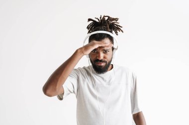 Kafası karışmış kulaklıklı Afro-Amerikan bir adamın beyaz arka planda izole edilmiş kameraya baktığı fotoğraf.