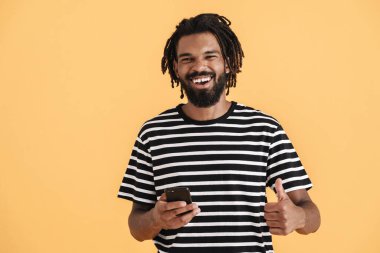 Neşeli Afrikalı Amerikalı adamın cep telefonu kullanırken ve sarı arka planda izole bir şekilde baş parmağını gösterirken fotoğrafı.