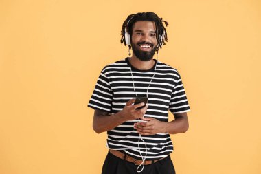 Sarı arka planda izole edilmiş cep telefonu ve kulaklık kullanan neşeli Afro-Amerikan adamın fotoğrafı.