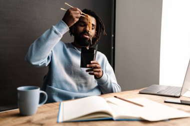 Sırıtkan Afrikalı genç adam masa başında çalışırken dışarıdan yemek alıyor.