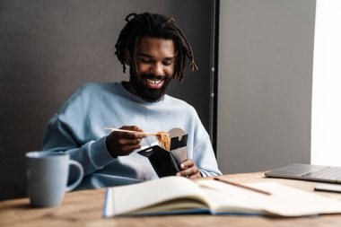 Sırıtkan Afrikalı genç adam masa başında çalışırken dışarıdan yemek alıyor.