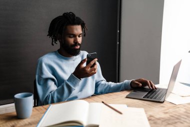 Zeki, ciddi bir Afrikalı adam masada oturuyor, dizüstü bilgisayar ve cep telefonuyla çalışıyor.