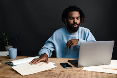 Yakışıklı Afrikalı bir adam kafeteryada oturmuş, dalgın dalgın bakıyor, iş planlarını düşünüyor, dizüstü bilgisayarla çalışıyor.