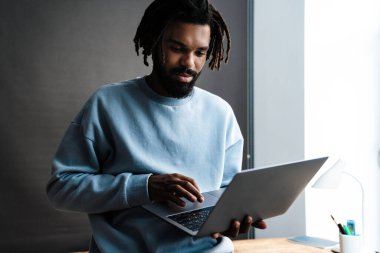 Kendine güveni olan Afro-Amerikan bir adam dizüstü bilgisayarla çalışırken bir masaya yaslanıyor.