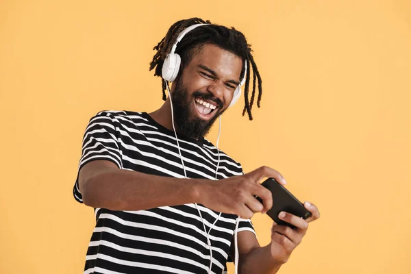 黄色の背景に隔離された携帯電話とヘッドフォンを使用して興奮したアフリカ系アメリカ人の男の写真 — ストック写真