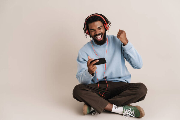 Excited african american guy in headphones using cellphone and making winner gesture isolated over white wall