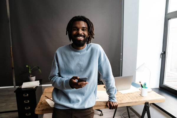 Smiling young african man holding mobile phone standing at the desk