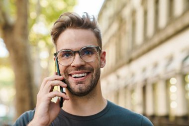 Gülümseyen çekici genç adam sokakta yürürken cep telefonuyla konuşuyor.