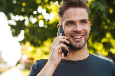 Gülümseyen çekici genç adam sokakta yürürken cep telefonuyla konuşuyor.