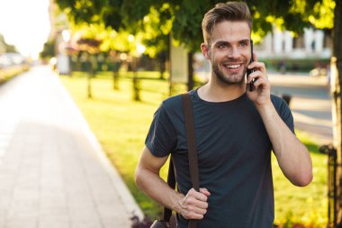 Gülümseyen çekici genç adam sokakta yürürken cep telefonuyla konuşuyor.