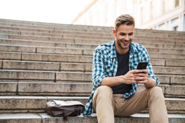 Dışarıda merdivenlerde otururken cep telefonu kullanan yakışıklı, gülümseyen genç adam.