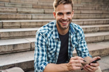 Dışarıda merdivenlerde otururken cep telefonu kullanan yakışıklı, gülümseyen genç adam.