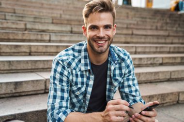 Dışarıda merdivenlerde otururken cep telefonu kullanan yakışıklı, gülümseyen genç adam.