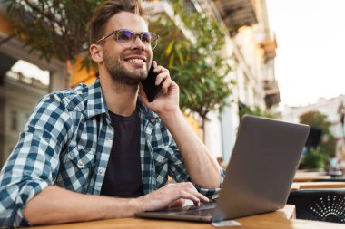 Kaldırımdaki kafede cep telefonu kullanırken dizüstü bilgisayarda çalışan gülümseyen genç adam.