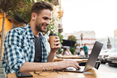 Kaldırımdaki kafede otururken dizüstü bilgisayarda çalışan mutlu genç adam.