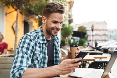 Kaldırımdaki kafede cep telefonu kullanırken dizüstü bilgisayarda çalışan gülümseyen genç adam.